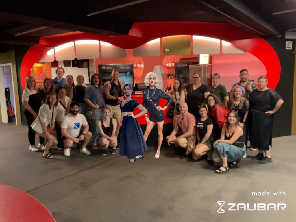 A group of people standing in front of Pepsi Boston Bar inside SchwuZ.  In their midst, in Augmented Reality, are the Berlin Drag Queens Jurassica Parka and Jacky-Oh Weinhaus. During this Berlin Pride event, people can take photos with Augmented reality.