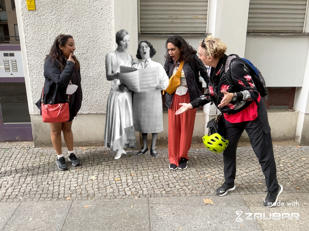 Three people standing around Claire Waldoff and another singer in Augmented Reality. During this Berlin Pride Event, people can discover queer history in Augmented Reality.